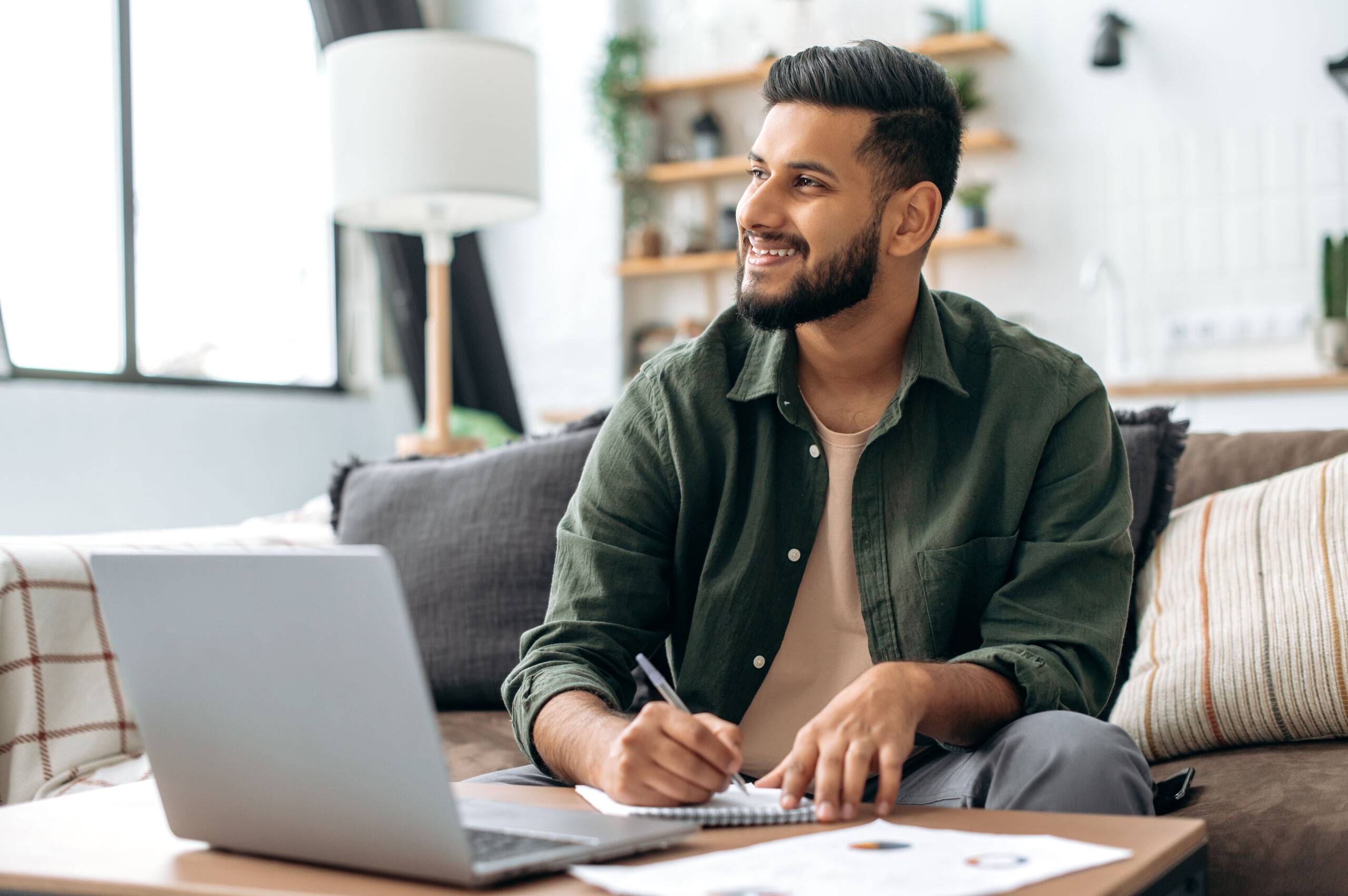 Positive handsome indian or arabian guy, student or freelancer, using laptop, working or studying remotely, listening to webinar, taking notes, homework while sitting at home on sofa in living room